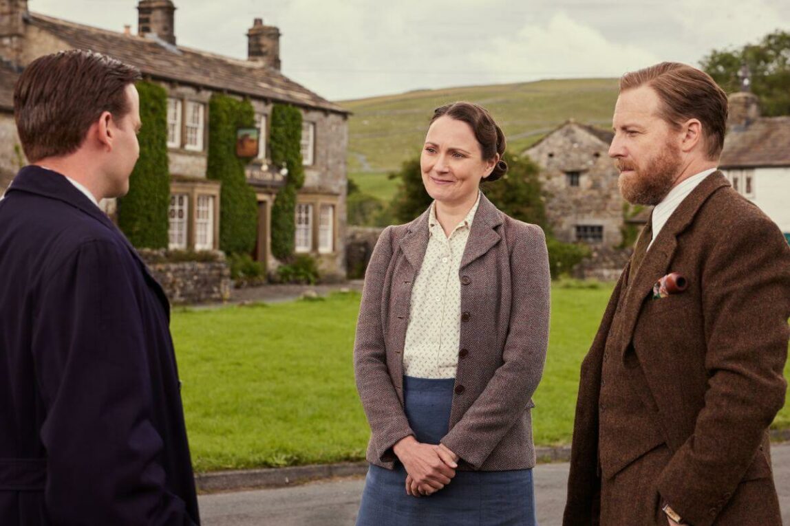 (L) James Herriot (M) Mrs Hall (R) Seigfried Farnon in All Creatures Great and Small Season 4 . The three are outside in front of a house.